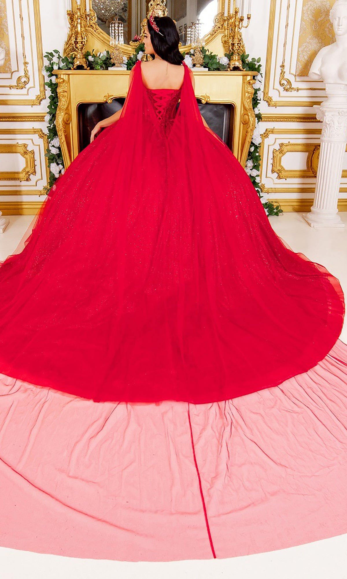 A woman stands in an ornate room in front of a golden fireplace adorned with floral decorations and a white statue nearby, wearing the Quinceanera Dress 8270J by Cinderella Couture—a long, flowing red glitter tulle ballgown with a train.